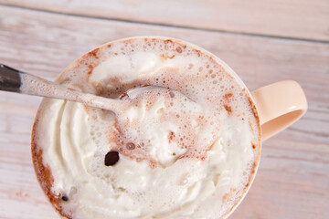 Cup of hot chocolate with whipped cream sprinkled with chocolate chips, being stirred with a spoon, on a wooden background