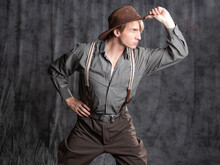 A Young Man In Retro Style, An Adventure Character. A Guy In A Hat And A Gray Shirt, Breeches With Suspenders.