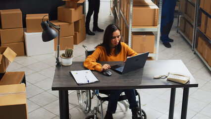 Poster - Young employee with impairment working in warehouse, doing desk work for stock logistics and inventory on laptop. Woman wheelchair user doing financial planning in disability friendly space.