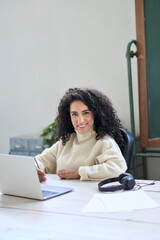 Wall Mural - Young happy hispanic business woman sitting at work desk with laptop. Smiling professional female worker sales or marketing manager looking at camera working or elearning on computer. Portrait