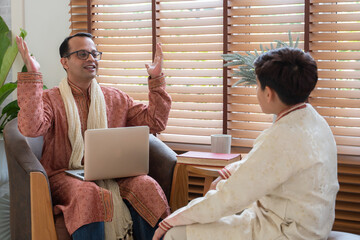 Indian father and handsome teenage son with traditional clothing sitting on sofa talking having conversation, smiling spending time together at home. Different aged generation communication in family