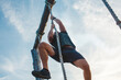 © Cavan Images - Below-view of a strong male pulling himself up a rope against blue sky