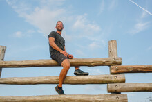 A Proud Smiling Man Sits High Atop A Tall Wood Fence Against Blue Sky