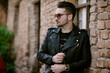 © Cavan Images - Trendy Man with sunglasses in a jacket standing by a brick wall.