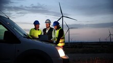 Team Work Of Three Renewable Energy Engineers With Helmets And Protection Equipment, Working And Planing At Night With Laptop Computer On Top Of The Car With A Wind Power Generators In The Background