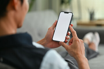 Young man resting on couch and checking social media on moblie phone. Over shoulder closeup view