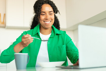 Wall Mural - Cheerful African American Woman Using Laptop Having Breakfast In Kitchen