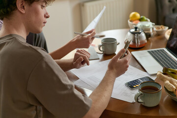 Cropped shot of serious thinking man paying utility bills payment online from home sitting in the kitchen with a laptop and paper invoices calculating and showing expenses to his wife on a smartphone.