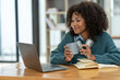 © crizzystudio - Young African businesswoman wearing headphones on her neck while looking at online shopping data Taking note of customer orders on a laptop while holding a mug of warm coffee to relax.