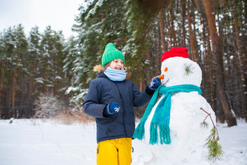 the boy made a snowman, attaches a carrot instead of a nose to the face of a snowman. happy child playing in snowy winter forest