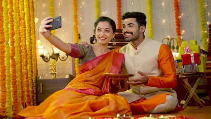 Wall Mural - Happy smiling indian couple taking selfie on mobile phone by holding flowers plate during diwali festival celebration at home - concept of togetherness, social media sharing and traditional culture.