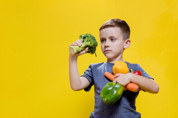 The boy holds fresh vegetables in his hands: broccoli, carrots and peppers on a yellow background. Vegan and healthy concepts.