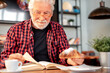 © luciano - Smiling senior bearded man sitting at cafe table reading a book while enjoying breakfast with espresso coffee cup and croissant. Elderly caucasian male in eyeglasses and checkered shirt