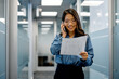 © Drazen - Happy Asian businesswoman talks on cell phone while reading documents in hallway.