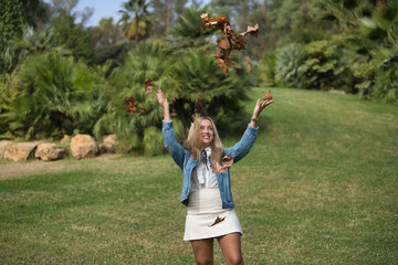 Young woman, beautiful, blonde and with green eyes, with denim jacket, white shirt and miniskirt, throwing a lot of dry leaves into the air, happy and cheerful. Concept autumn, leaves, happiness.