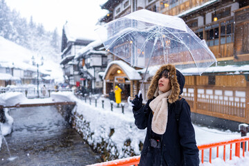 Young Asian woman tourist holding umbrella walking down footpath at onsen area Ginzan Onsen in Yamagata prefecture, Japan in snowy day. Attractive girl travel local village landmark on winter vacation