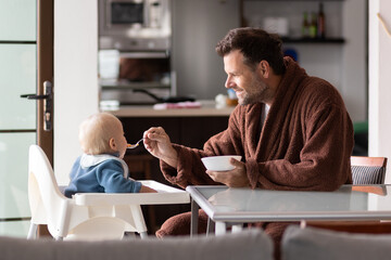 Wall Mural - Father wearing bathrope spoon feeding hir infant baby boy child sitting in high chair at the dining table in kitchen at home in the morning.