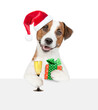 Happy Jack russell terrier puppy wearing red santa hat holds gift box and glass of champagne and looks above  empty white banner. isolated on white background