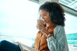 © Nina Lawrenson/peopleimages.com - Airport, coffee and black woman reading a book by window waiting for global flight, departure and travel. International transport, lobby and girl with headphones for music, drinking beverage and read
