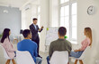 © Studio Romantic - Businessman making presentation to his colleagues during meeting or in-house business training. Young man at board in office speaks to his business team sitting on chairs in front of him.