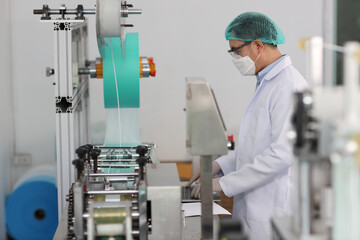 Poster - Worker man in personal protective equipment or PPE inspecting quality of mask and medical face mask production line in factory, manufacturing industry and factory concept.