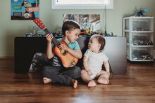 Baby Sister And Big Brother With Guitar Sitting On Hardwood Floor