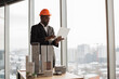 © sofiko14 - Handsome chief architect african man in business suit and protective helmet with laptop pc working with city buildings model as urban planner at office interior with panoramic city view.
