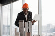 © sofiko14 - Focused african inspector using laptop inspecting skyscraper model in architectural modern office with panoramic city view on architectural bureau. Handsome man wearing suit and hardhat.