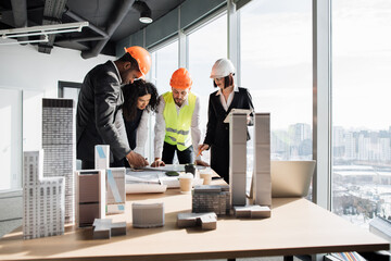 Multiracial coworkers builders and architects standing near table with blueprints, gadgets and design of buildings residential project maquette. Business people working on construction project