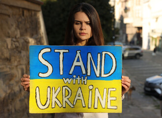 Poster - Sad woman holding poster in colors of national flag and words Stand with Ukraine on city street. Space for text