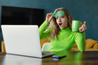 A young woman in green clover-shaped glasses looks in surprise at a laptop screen at a table in the room. Holds a cup with a drink for St. Patrick's Day
