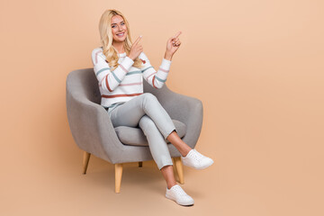 Poster - Full length photo of attractive lady blonde hair wear striped sweater sit comfort armchair point fingers mockup isolated on beige color background