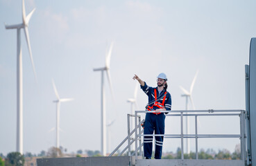 Wall Mural - Engineer man or technician worker stand on base of windmill or wind turbine and point to left side and stay in front of windmill cluster in background with blue sky.