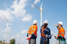 Three Engineers Or Technician Workers Work Together In Front Of Cluster Of Windmill Or Wind Turbine With Blue Sky And Some Cloud In Their Workplace.