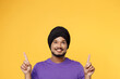 Happy devotee Sikh Indian man ties his traditional turban dastar wear purple t-shirt point index finger overhead on area isolated on plain yellow background studio portrait. People lifestyle concept.