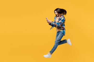 Wall Mural - Full body side view happy young smiling woman wear blue shirt beige t-shirt jump high hold in hand use mobile cell phon isolated on plain yellow background studio portrait. People lifestyle concept.