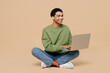 Full body young man of African American ethnicity wearing green sweatshirt sitting hold use work on laptop pc computer isolated on plain pastel light beige background studio. People lifestyle concept.