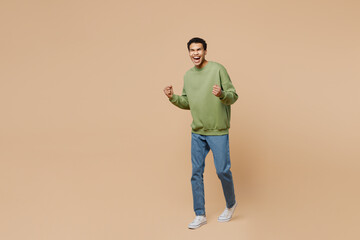 Full body excited happy young man of African American ethnicity wear green sweatshirt walk so winner gesture isolated on plain pastel light beige background studio portrait. People lifestyle concept.