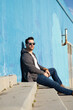 © Cavan Images - Portrait of stylish man sitting on stairs leaning on a blue wall while looking camera