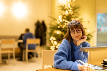 Wall Mural - Portrait of young adult woman sitting at modern coffee shop in yellow tones and Cristmas tree on background. Concept of remote creative work online