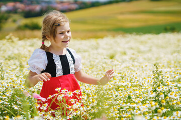 Wall Mural - Little preschool girl in daisy flower field. Cute happy child in red riding hood dress play outdoor on blossom flowering meadow with daisies. Leisure activity in nature with children.