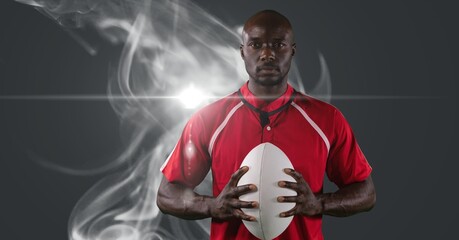 Wall Mural - African american male rugby player holding ball against smoke and light spot on grey background