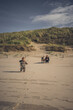 Kleinkind läuft am Stand durch den Sand und hat Spaß, im Hintergrund beobachtet Mutter mit Hund das Mädchen im bunten Overall