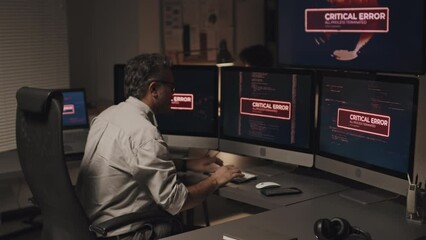 Rear view of mature programmer fighting with hacking of system, he sitting at table and typing security codes to solve the problem