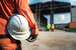 © Nattawit - A construction worker is holding white safety helmet or hardhat with construction work site as background. Safe working practice in the industrial scene. Generative Ai image.