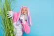 Photo of tricky cunning retired man wear pink hare pajama hiding green plant looking finger mouth empty space isolated blue color background