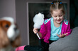 Portrait of smiling little girl holding candyfloss, looking at sticky fingers hand, sitting on sofa near dog at home.