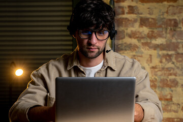 A focused young businessman in glasses and headphones, conducting a video call with customers on a laptop. A focused man with glasses reads an online lecture of an educational class.