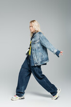 Full Length Of Young Blonde Woman In Blue Denim Outfit And Stylish Sneakers Walking On Grey.