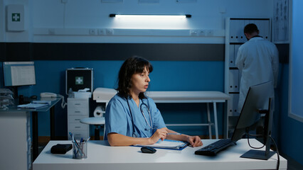 Wall Mural - Physician nurse with blue uniform and stethoscope analyzing patient disease report working at health care treatment late at night in hospital office. Assistant checking medical expertise on computer